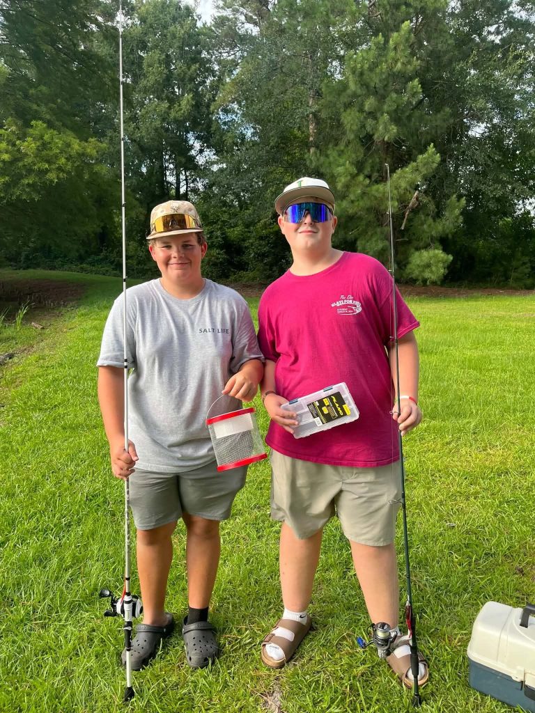 Two boys stand on grass holding fishing rods and supplies, dressed casually in shorts, t-shirts, and hats, with trees and greenery in the background.