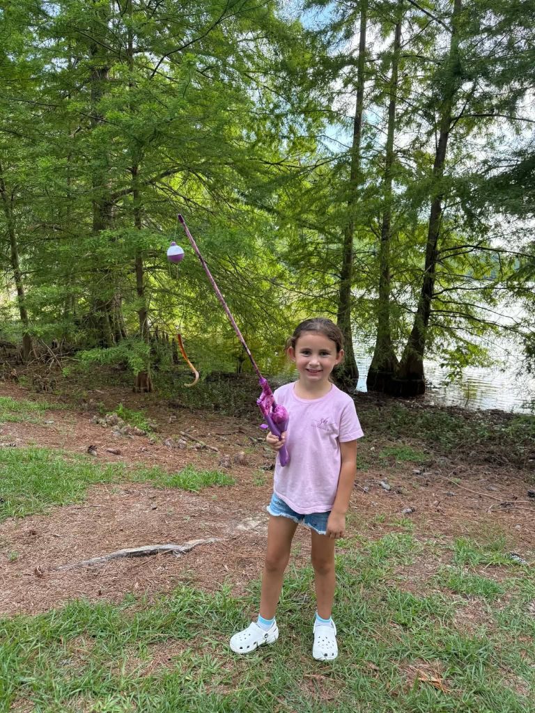 Young girl in pink shirt and shorts holding a fishing pole outdoors near a lake, surrounded by green trees and grass.