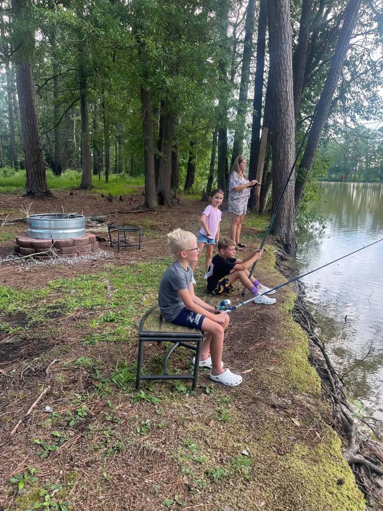 Three children and one adult fish by a wooded lakeside, with fishing rods, near a makeshift fire pit and surrounded by tall trees.