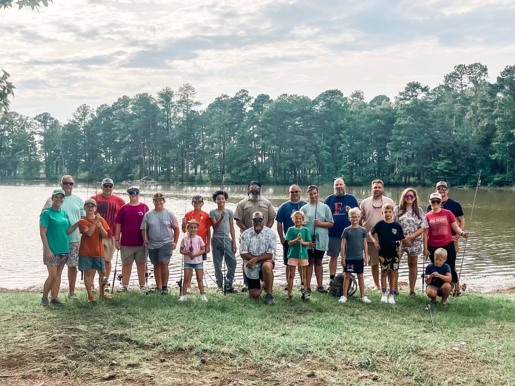 A group of people, including children and adults, pose together outdoors near a lake with fishing poles and trees in the background.