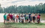A group of people, including children and adults, pose together outdoors near a lake with fishing poles and trees in the background.