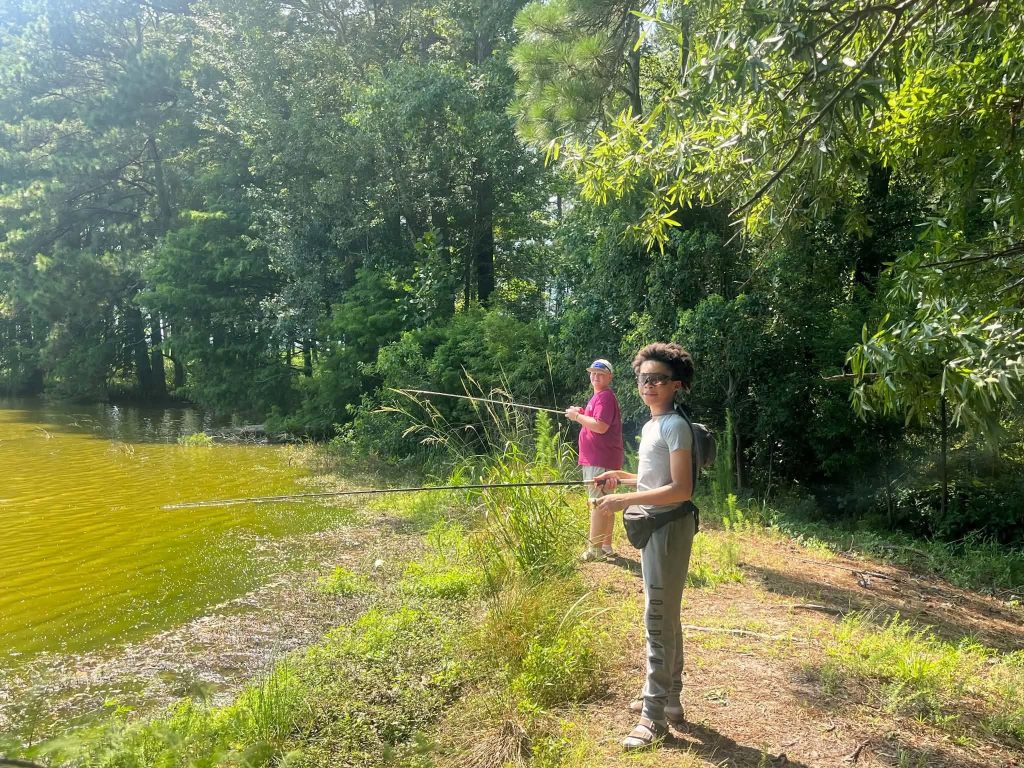 Two people fishing by a lake shore, surrounded by green trees and foliage, under bright sunlight.