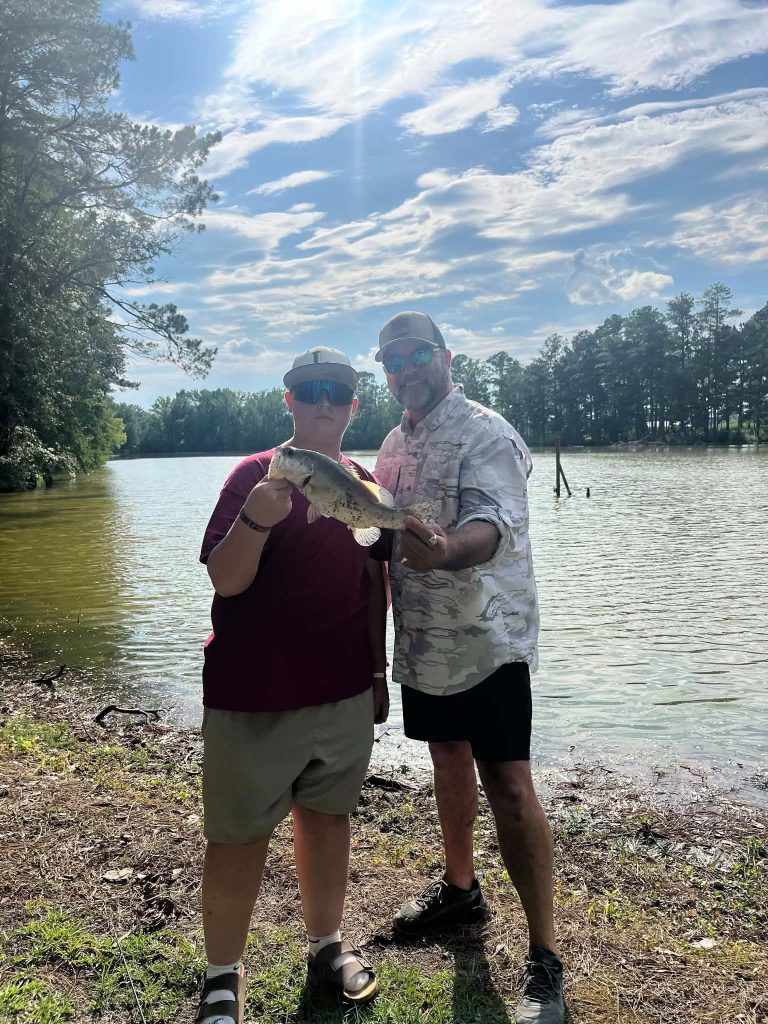Two people stand by a lake, smiling and holding a large fish they caught, surrounded by trees and under a partly cloudy sky.