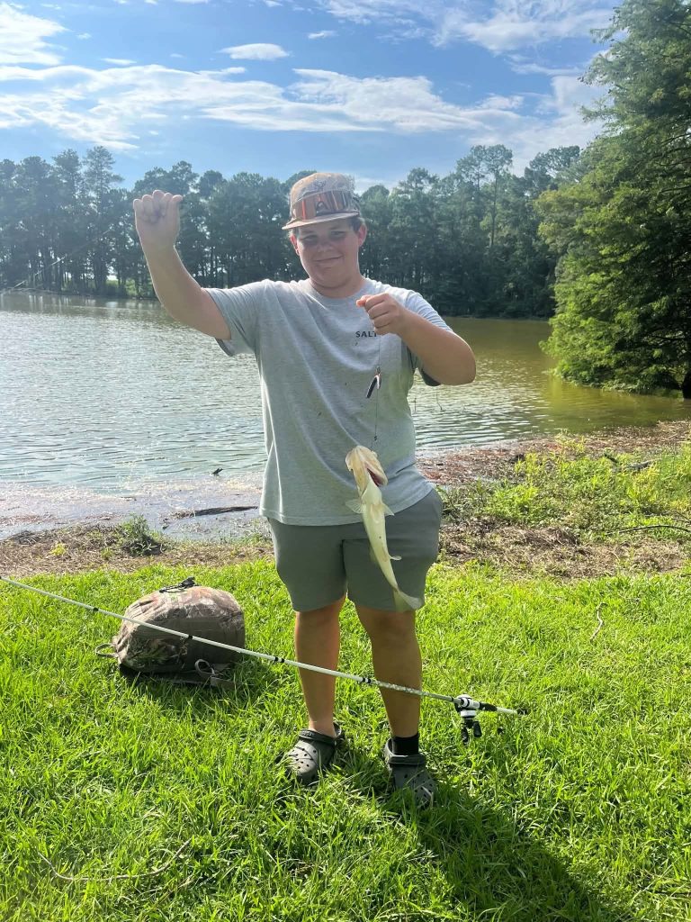 Person standing on grassy bank by a lake, holding a fishing rod and displaying a freshly caught fish, with trees and blue sky in the background.