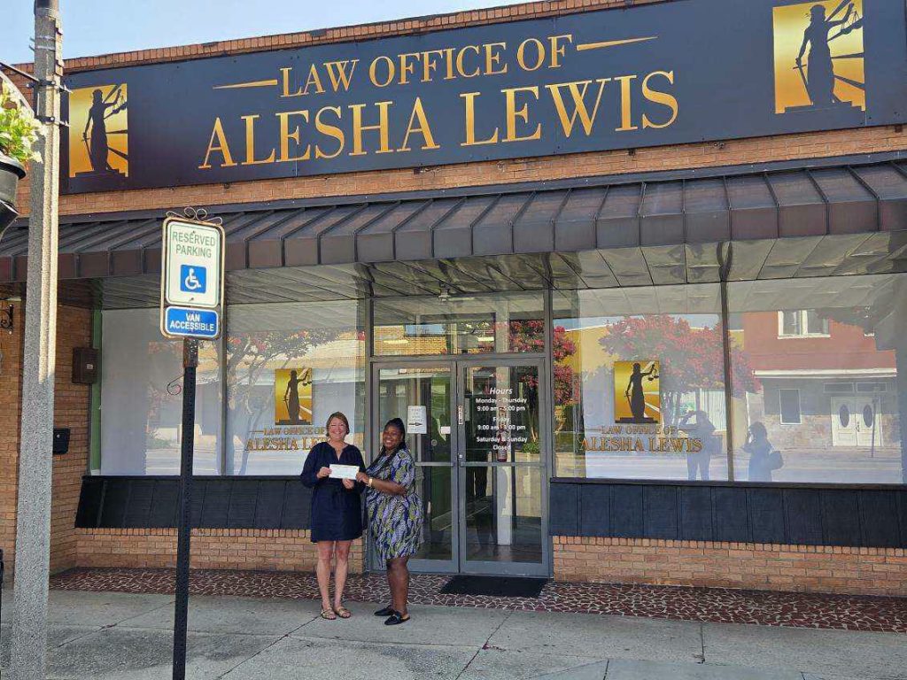 Two women stand smiling in front of the Law Office of Alesha Lewis, next to a reserved parking sign for van accessibility.