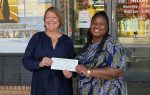 Two women smiling and holding a large check outside an office building with business hours and a justice-themed sign visible in the background.