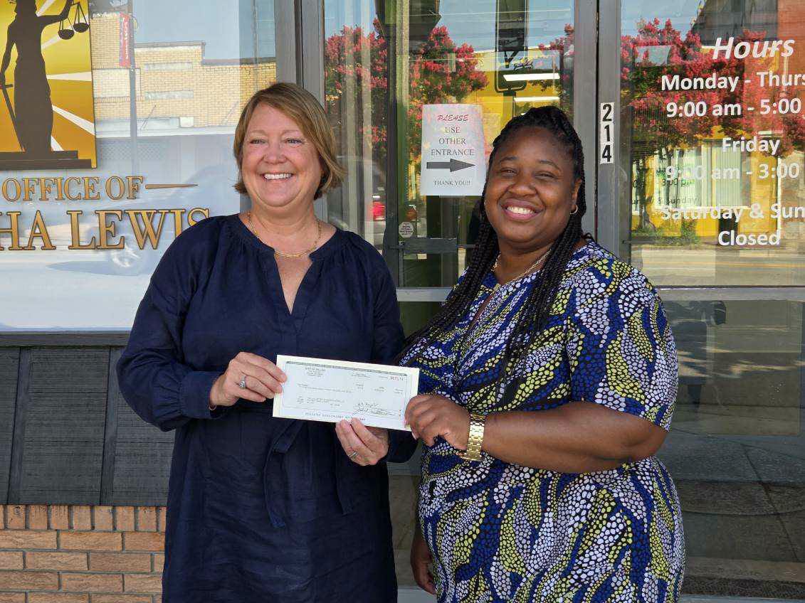 Two women smiling and holding a large check outside an office building with business hours and a justice-themed sign visible in the background.