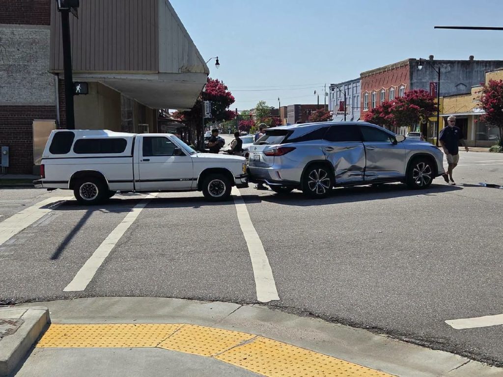 A white pickup truck and a silver SUV involved in a collision at an intersection, with visible damage and people standing nearby on a sunny day.