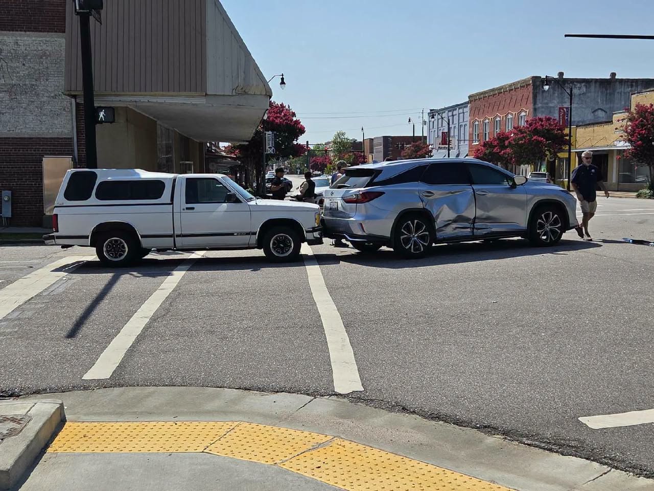 A white pickup truck and a silver SUV involved in a collision at an intersection, with visible damage and people standing nearby on a sunny day.