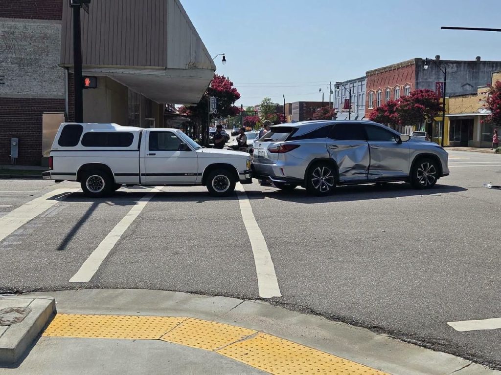 Two vehicles, a white truck and a silver SUV with visible side damage, are involved in a collision at an intersection in a small downtown area.