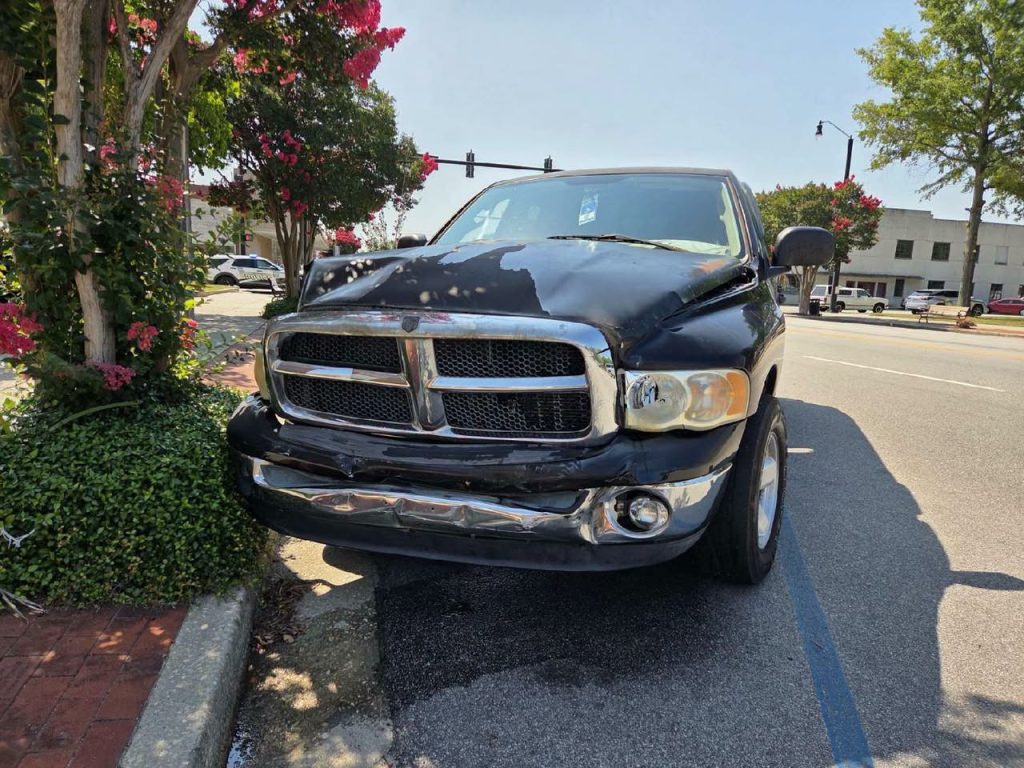 A black pickup truck with heavy front-end damage is parked partially on a curb beside some bushes and trees on a sunny street.