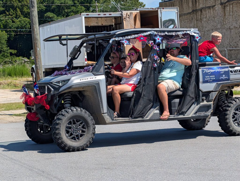 A decorated off-road vehicle with people inside, including a child riding on the back, drives in a parade. Patriotic decorations adorn the vehicle.
