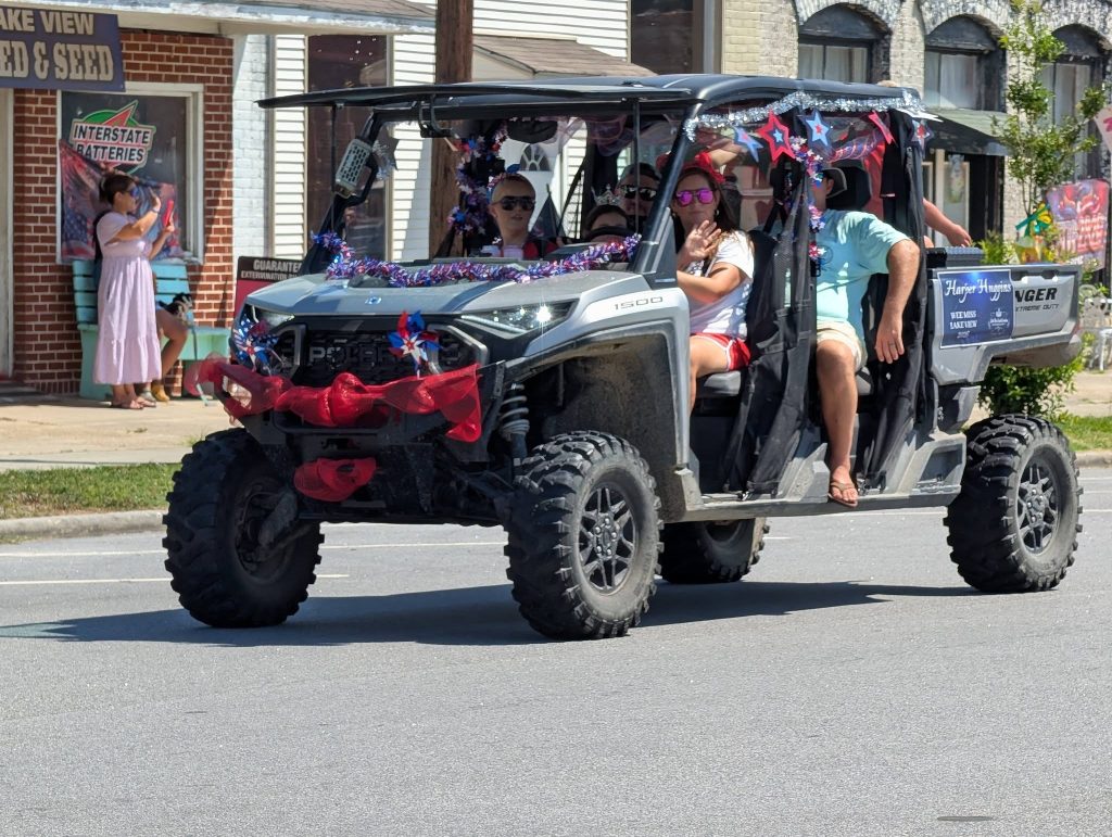 A decorated off-road vehicle with people inside drives in a parade on a sunny day, passing a seed store with spectators on the sidewalk.