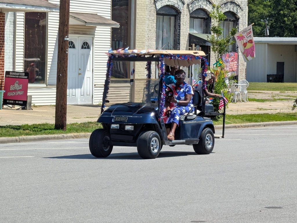 A decorated golf cart drives in a parade; two people ride, wearing patriotic clothing and holding small American flags, with festive decorations on the vehicle.