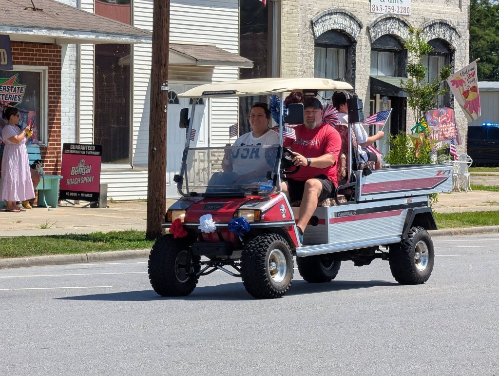 A group of people riding a decorated golf cart with American flags during a parade, on a sunny day in a small town.