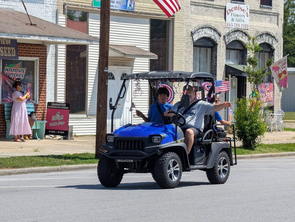 A blue utility vehicle with people inside drives down a street with American flags, passing storefronts and a woman in a pink dress standing on the sidewalk.