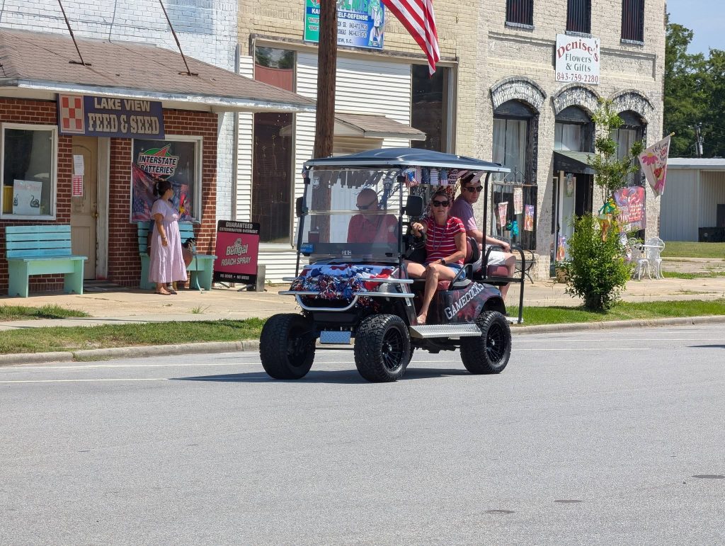 People riding a decorated golf cart past small-town shops, with an American flag and a woman in a pink dress standing on the sidewalk.