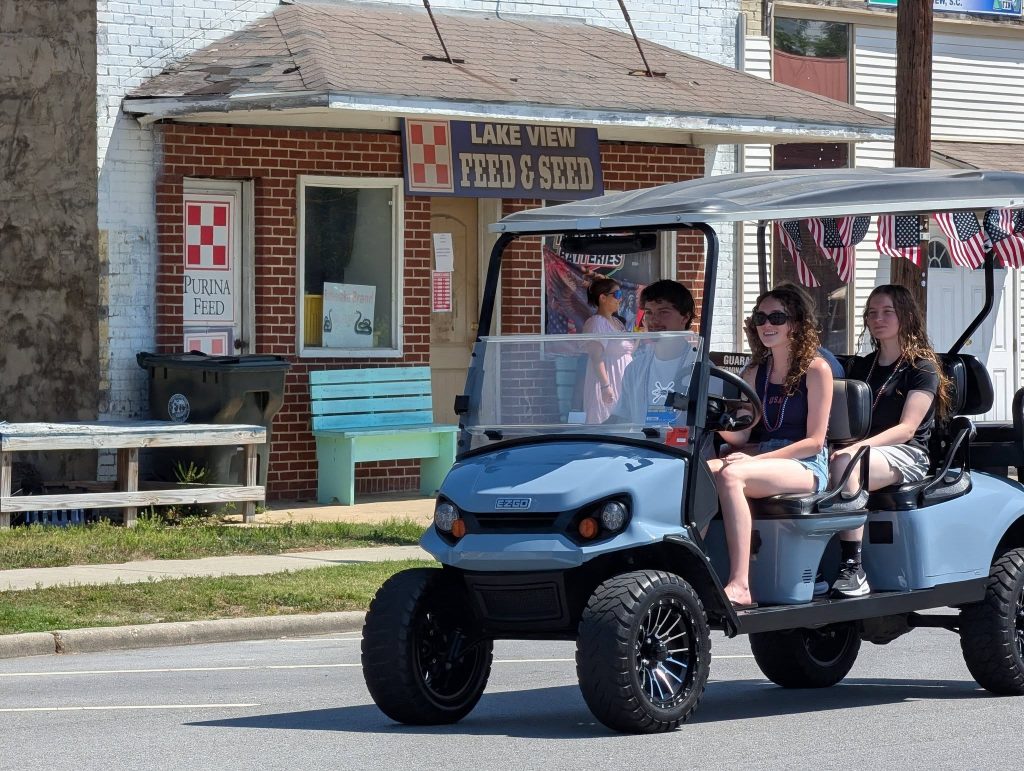 Three young people ride a blue golf cart decorated with American flags, passing a brick feed store on a sunny street.
