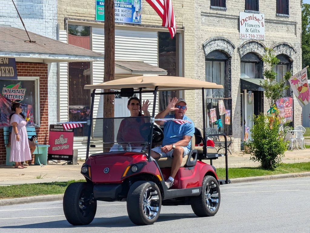 Two people wave from a red golf cart driving down a sunny street, with shops and American flags visible in the background.