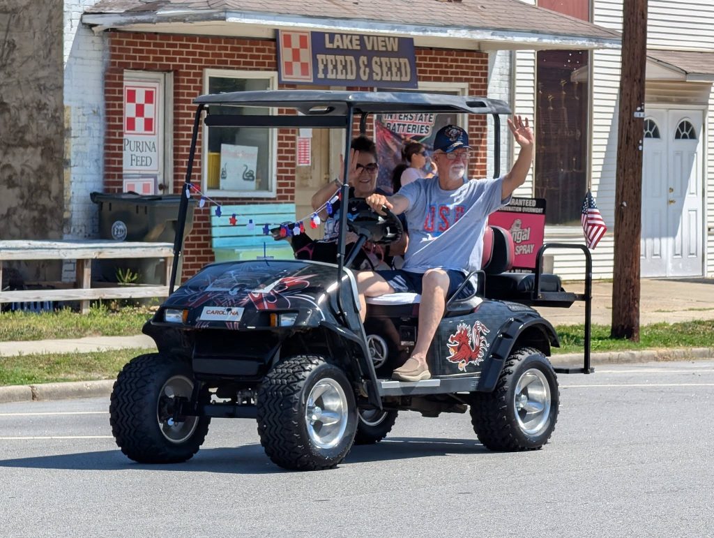 Two people ride a decorated golf cart with American flags and patriotic colors, driving past a feed store in a small-town street parade.