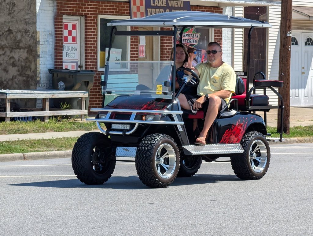 Two people ride a custom black golf cart with red flame decals on a street in front of a brick building and storefronts on a sunny day.