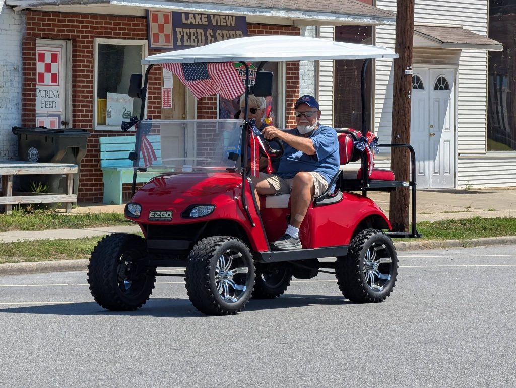 A man drives a bright red golf cart decorated with American flags down a street, passing by a brick storefront labeled "Lake View Feed & Seed."
