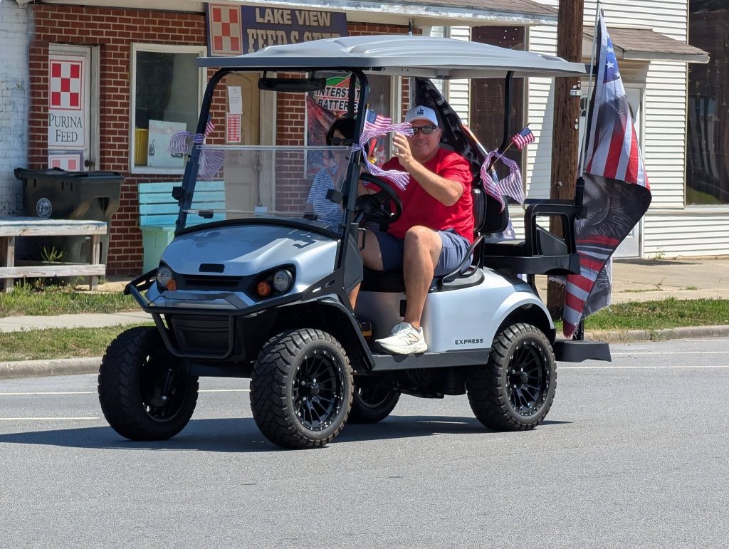 Man in sunglasses and red shirt drives a decorated golf cart with American flags on a street in front of a store.