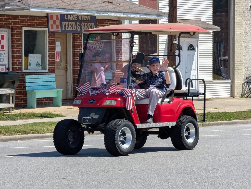 elderly man waves from a red golf cart decorated with small American flags, driving down a street in front of a feed and seed store.