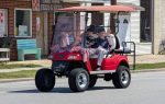 elderly man waves from a red golf cart decorated with small American flags, driving down a street in front of a feed and seed store.