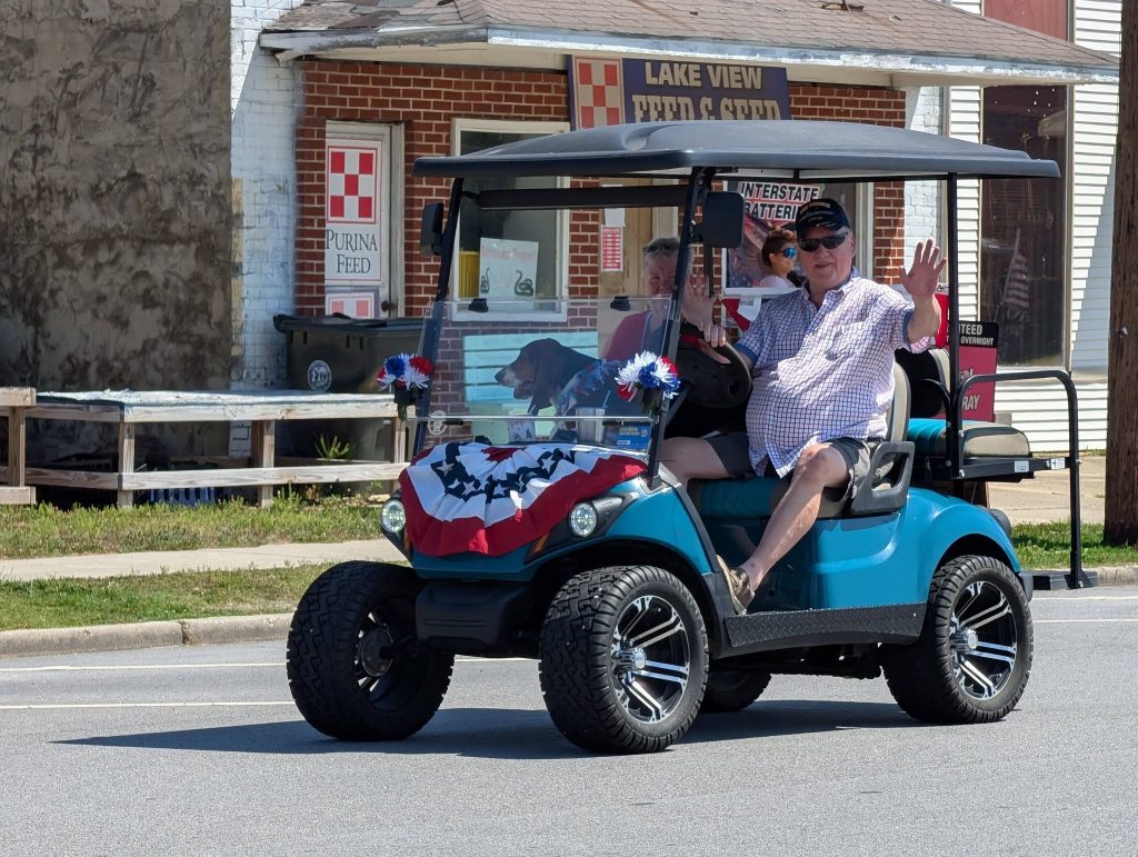 A man waves from a decorated blue golf cart with a dog inside, driving on a street in front of a feed and seed store.