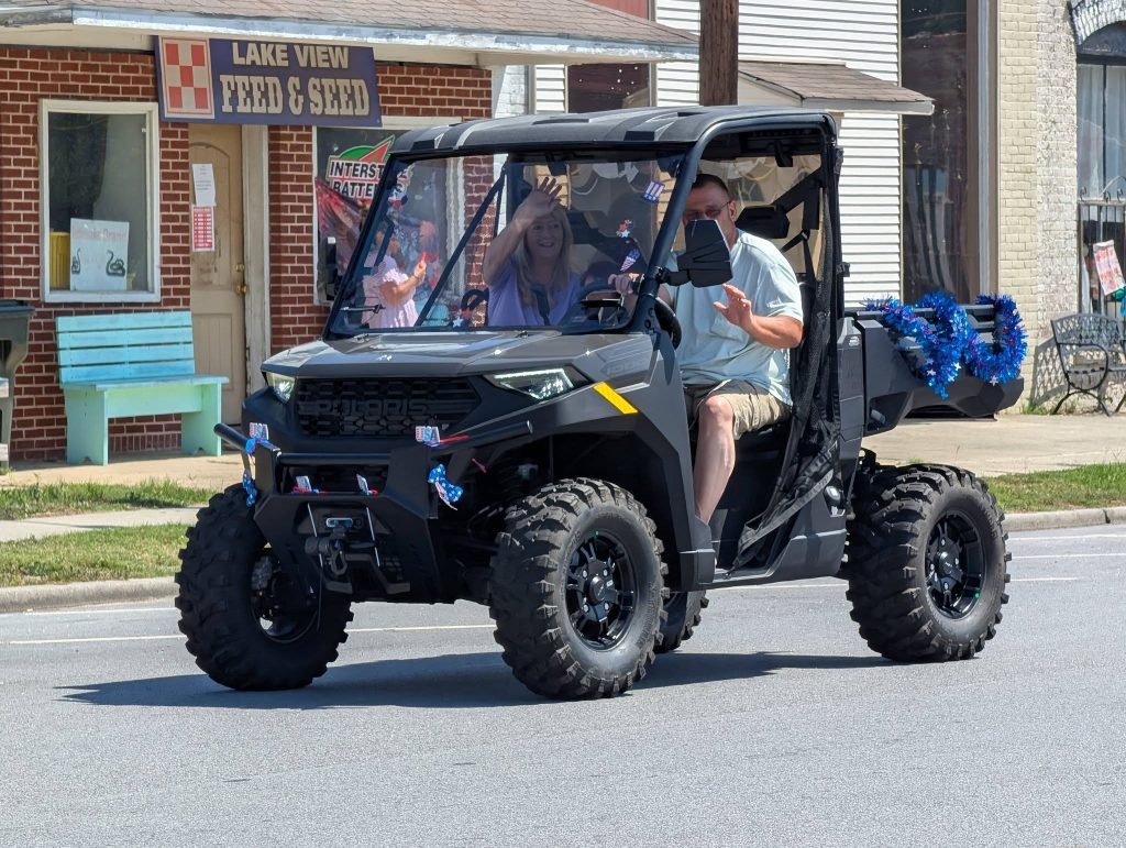 Man and woman ride in a decorated black ATV on a sunny street, waving and smiling, passing a feed and seed store in a small-town setting.