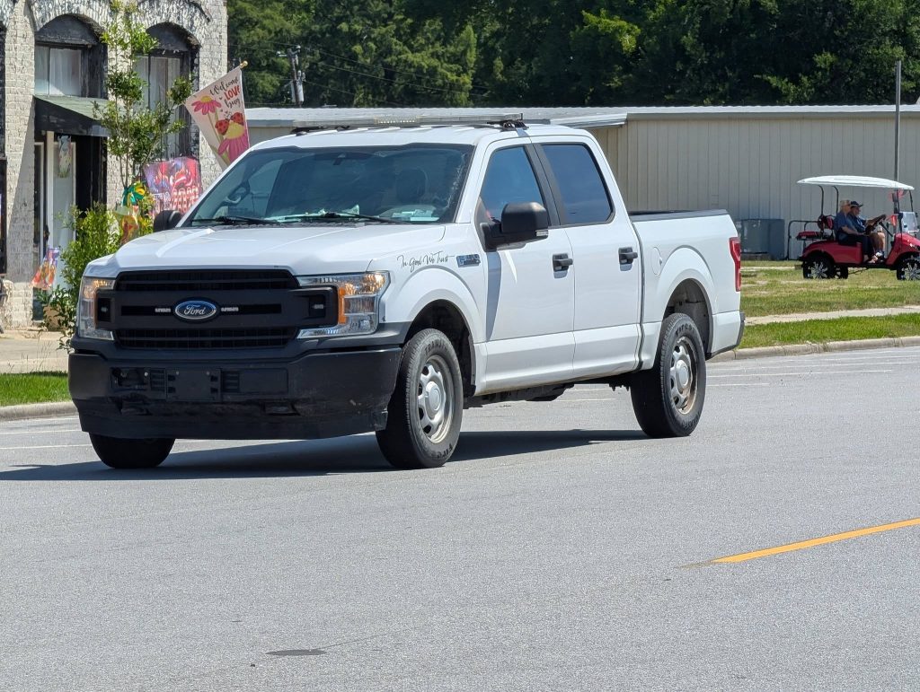 A white Ford F-150 pickup truck driving on a sunlit street, with buildings and a red golf cart in the background.