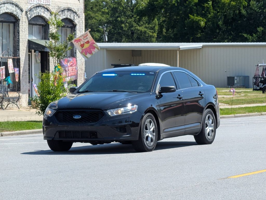 A black unmarked Ford police car with blue emergency lights is driving on a sunny street near shops and a bench.