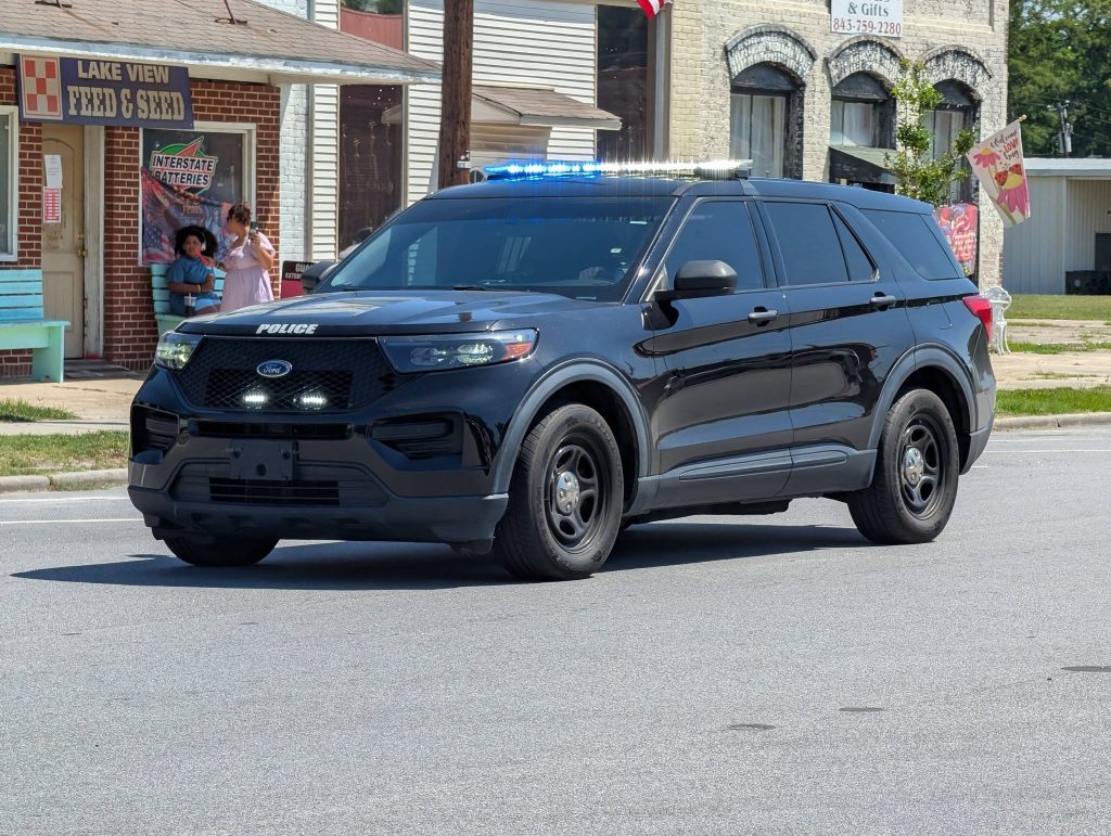 A black police SUV with blue lights on, driving on a street in front of shops. Two people stand on the sidewalk nearby.