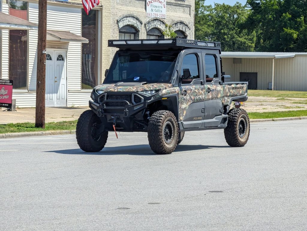 A camouflaged Polaris Ranger utility vehicle with off-road tires is parked on a street in front of older commercial buildings.