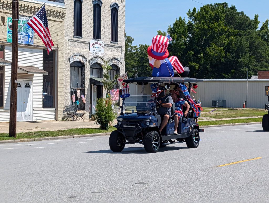 People riding a decorated golf cart with a large Uncle Sam figure and American flags, passing by storefronts during a patriotic parade on a sunny day.