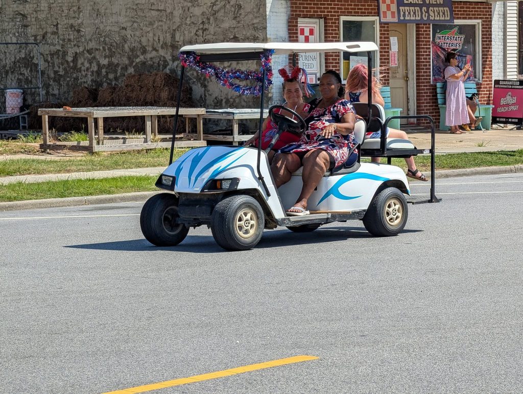 Two women ride a decorated golf cart in a parade. Another woman stands on the street, and a brick store is visible in the background.