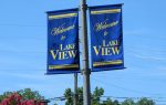 Two blue banners on a pole read "Welcome to Lake View" in yellow letters, with trees and power lines in the background under a clear blue sky.
