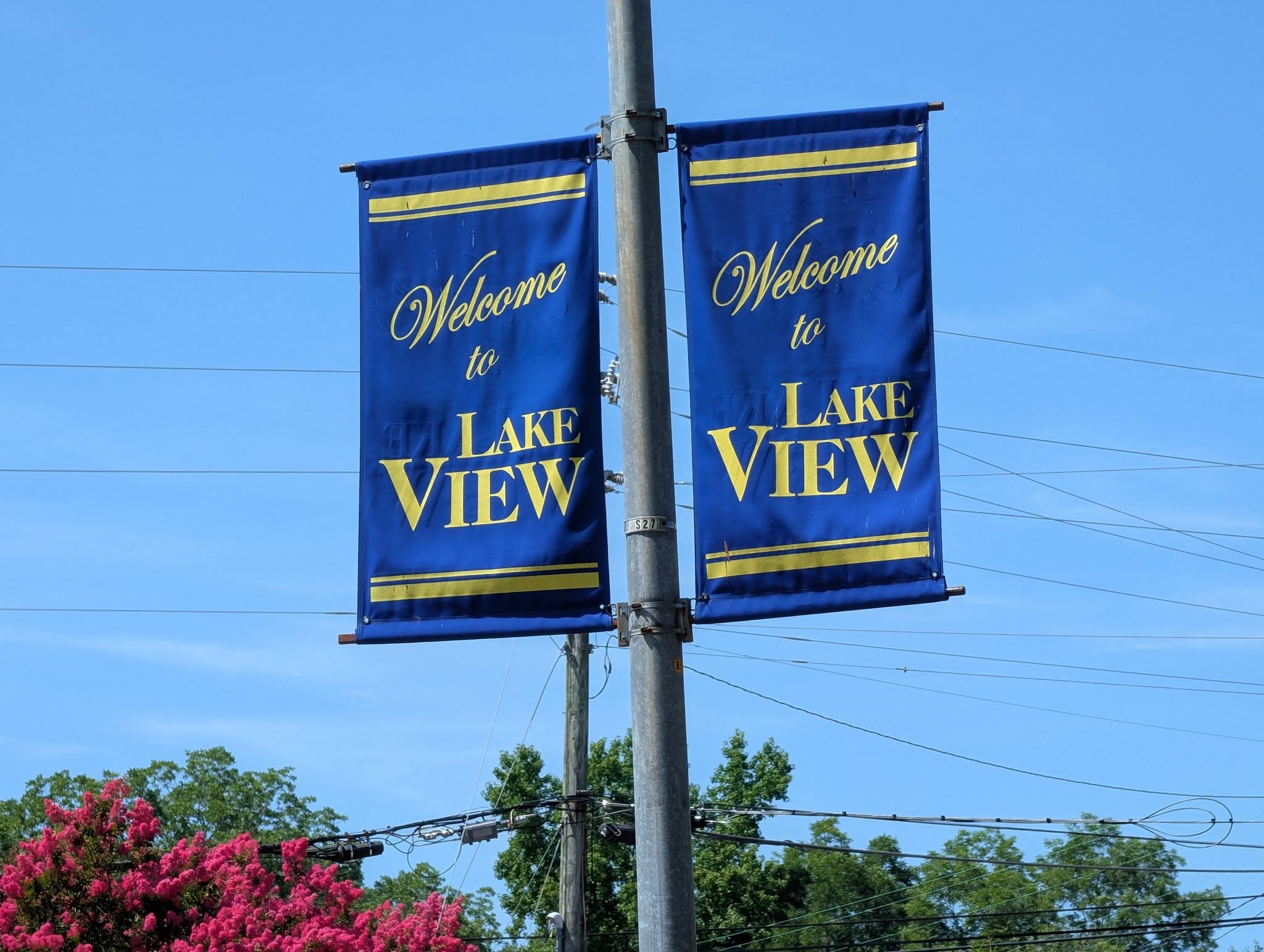 Two blue banners on a pole read "Welcome to Lake View" in yellow letters, with trees and power lines in the background under a clear blue sky.