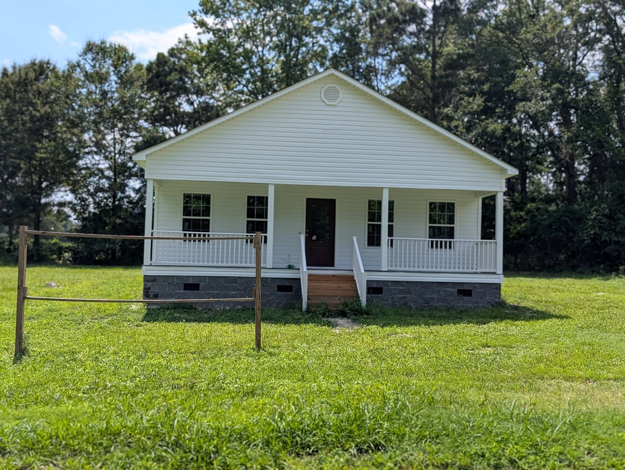 A small white house with a porch and steps, surrounded by green grass and trees, under a partly cloudy sky.
