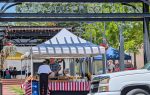 Outdoor market scene at Main Street Commons with vendor tents, people shopping, and a white truck parked in front. Striped tablecloth and hanging flowers visible.