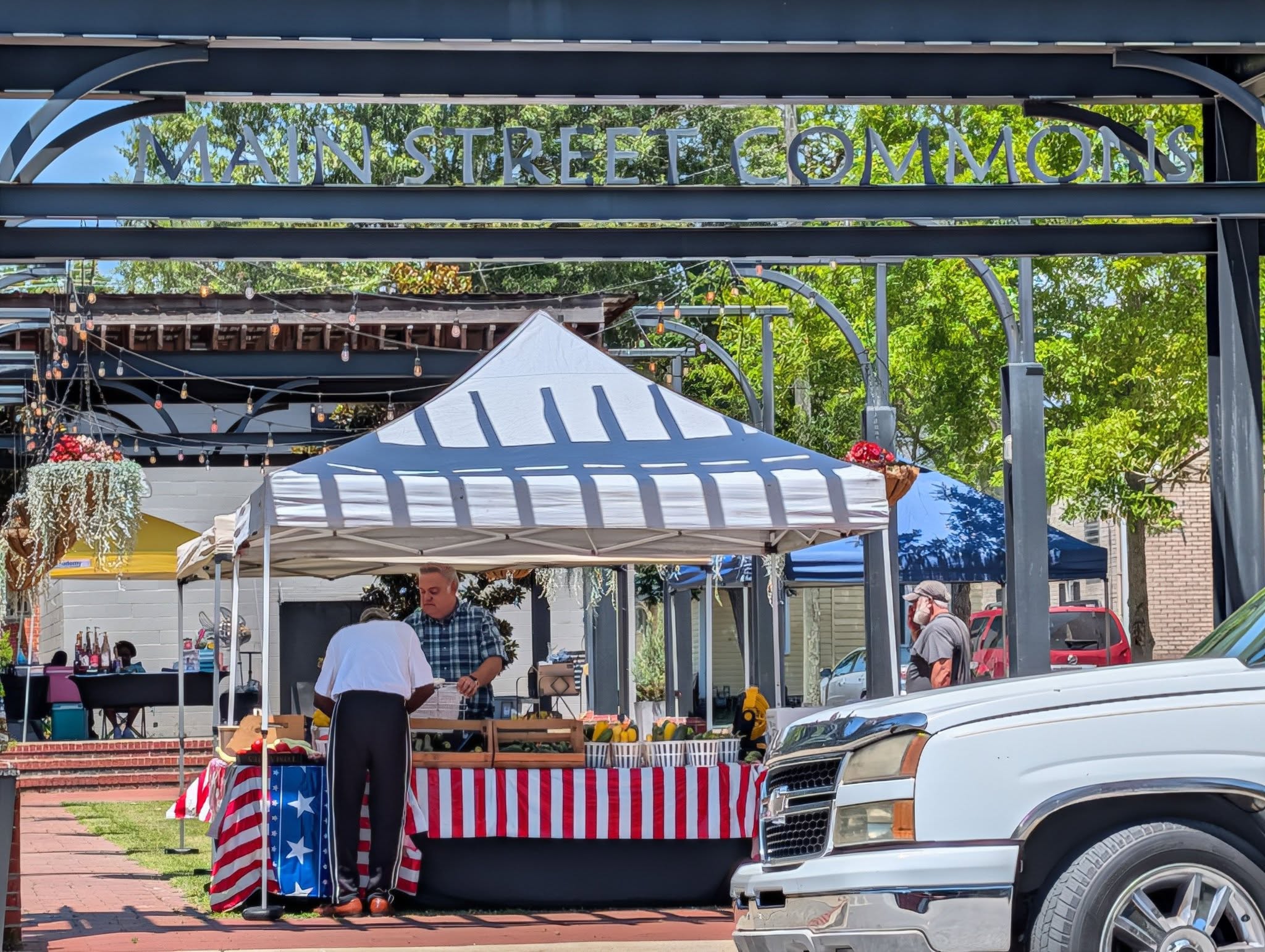 Outdoor market scene at Main Street Commons with vendor tents, people shopping, and a white truck parked in front. Striped tablecloth and hanging flowers visible.