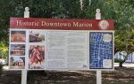 Signboard for Historic Downtown Marion with historical text, photos of landmarks, and a map displayed outdoors near trees and grass.