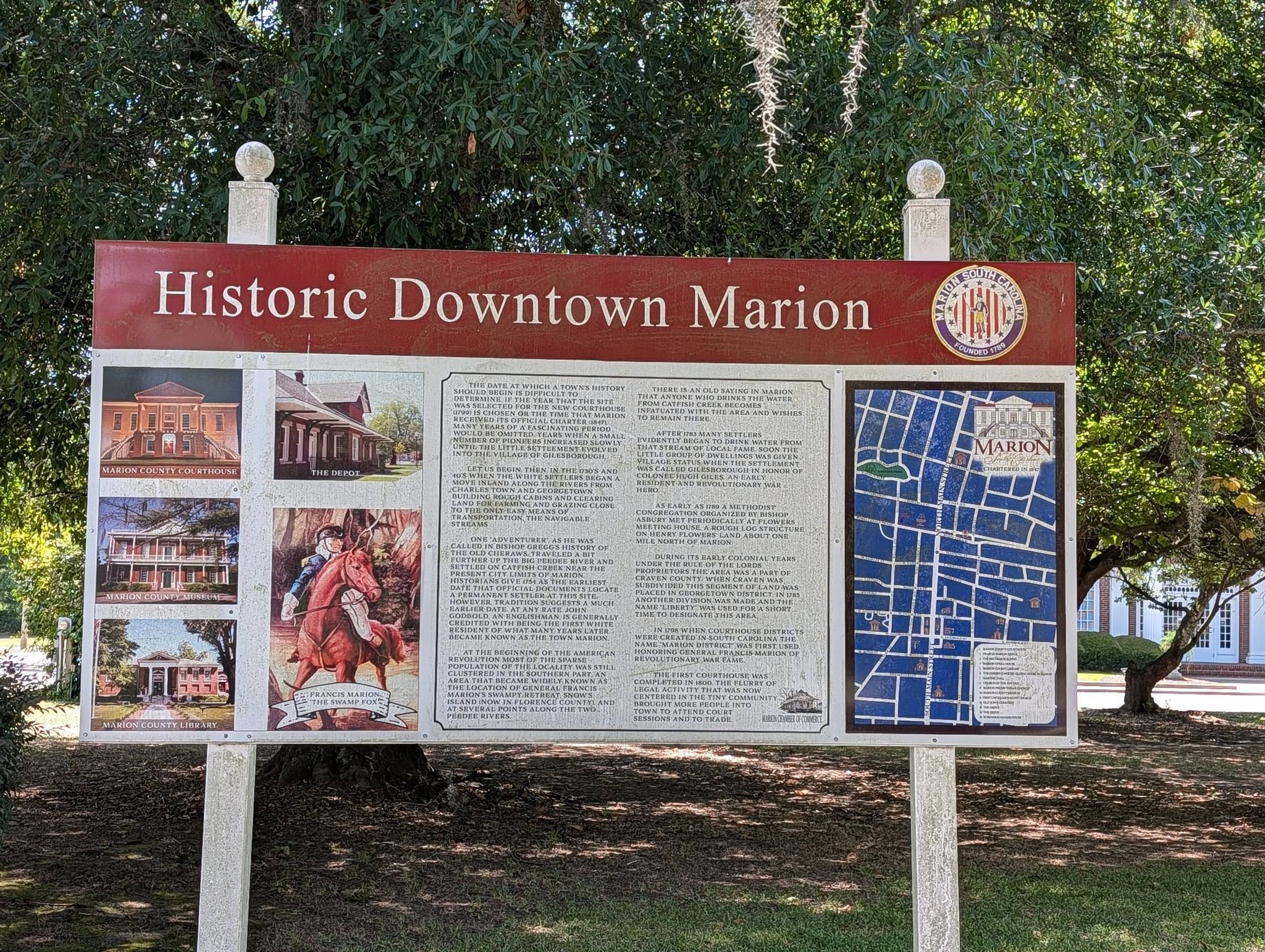Signboard for Historic Downtown Marion with historical text, photos of landmarks, and a map displayed outdoors near trees and grass.