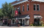 A red-brick corner building houses FoxySteel Boutique, with large display windows and street signs. Green trees and American flags line the sidewalk on a sunny day.