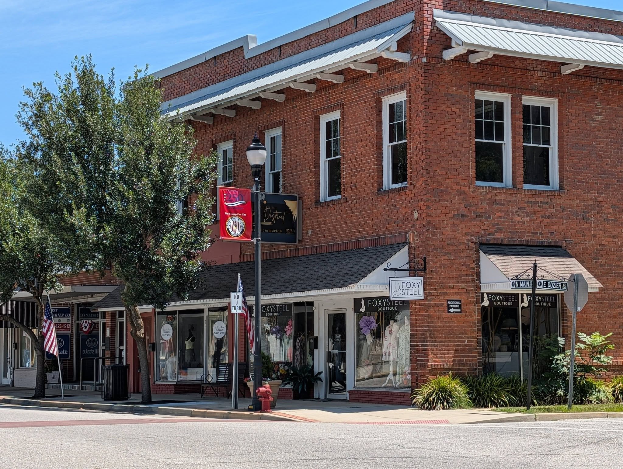 A red-brick corner building houses FoxySteel Boutique, with large display windows and street signs. Green trees and American flags line the sidewalk on a sunny day.