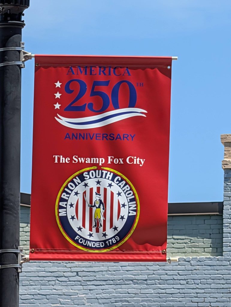 Red banner celebrating America’s 250th anniversary, featuring “The Swamp Fox City” and the seal of Marion, South Carolina, founded 1789, against a blue sky background.