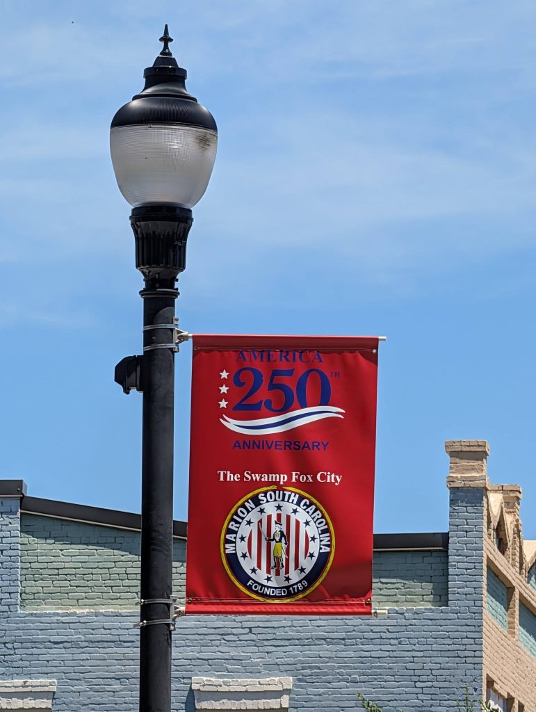 A streetlamp with a red American 250th anniversary banner for Marion, South Carolina, reading "The Swamp Fox City," and buildings in the background.