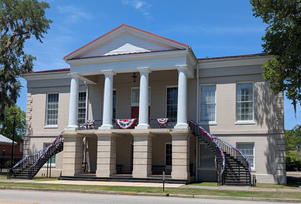 Historic two-story courthouse with white columns, two staircases, and patriotic red, white, and blue bunting decorations, set against a bright blue sky.