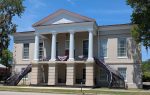 Historic two-story courthouse with white columns, two staircases, and patriotic red, white, and blue bunting decorations, set against a bright blue sky.